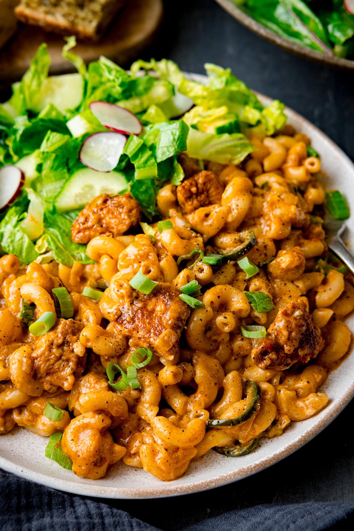 A plate of Buffalo Chicken Macaroni Cheese with seasoned meat, topped with spring onions, served alongside a fresh green salad with cucumber and radish slices.