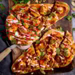 Flatbread pizzas topped with grilled chicken tikka, tomato sauce, coriander, sliced almonds, and a drizzle of white sauce. This chicken tikka naan pizza is served on a wooden board with a pizza cutter.