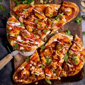 Flatbread pizzas topped with grilled chicken tikka, tomato sauce, coriander, sliced almonds, and a drizzle of white sauce. This chicken tikka naan pizza is served on a wooden board with a pizza cutter.