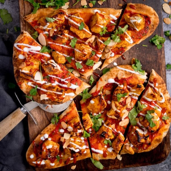Flatbread pizzas topped with grilled chicken tikka, tomato sauce, coriander, sliced almonds, and a drizzle of white sauce. This chicken tikka naan pizza is served on a wooden board with a pizza cutter.