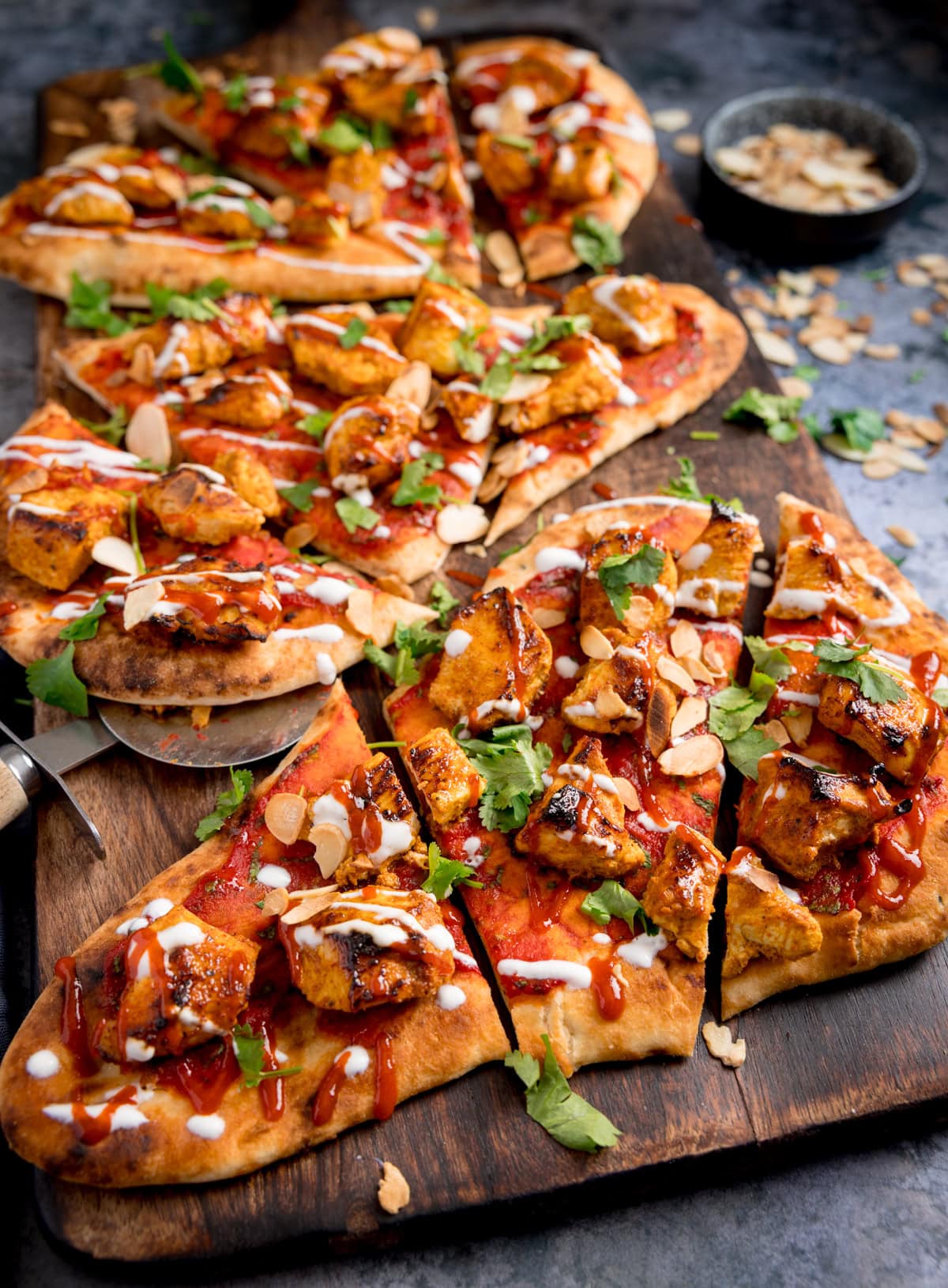 Several slices of Chicken tikka pizza topped with grilled chicken, fresh coriander, sliced almonds, and drizzles of red and white sauce, served on a wooden board.