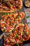 Sliced chicken tikka naan pizza topped with coriander, flaked almonds, and drizzled with sauces on a wooden board. A bowl of flaked almonds sits nearby.