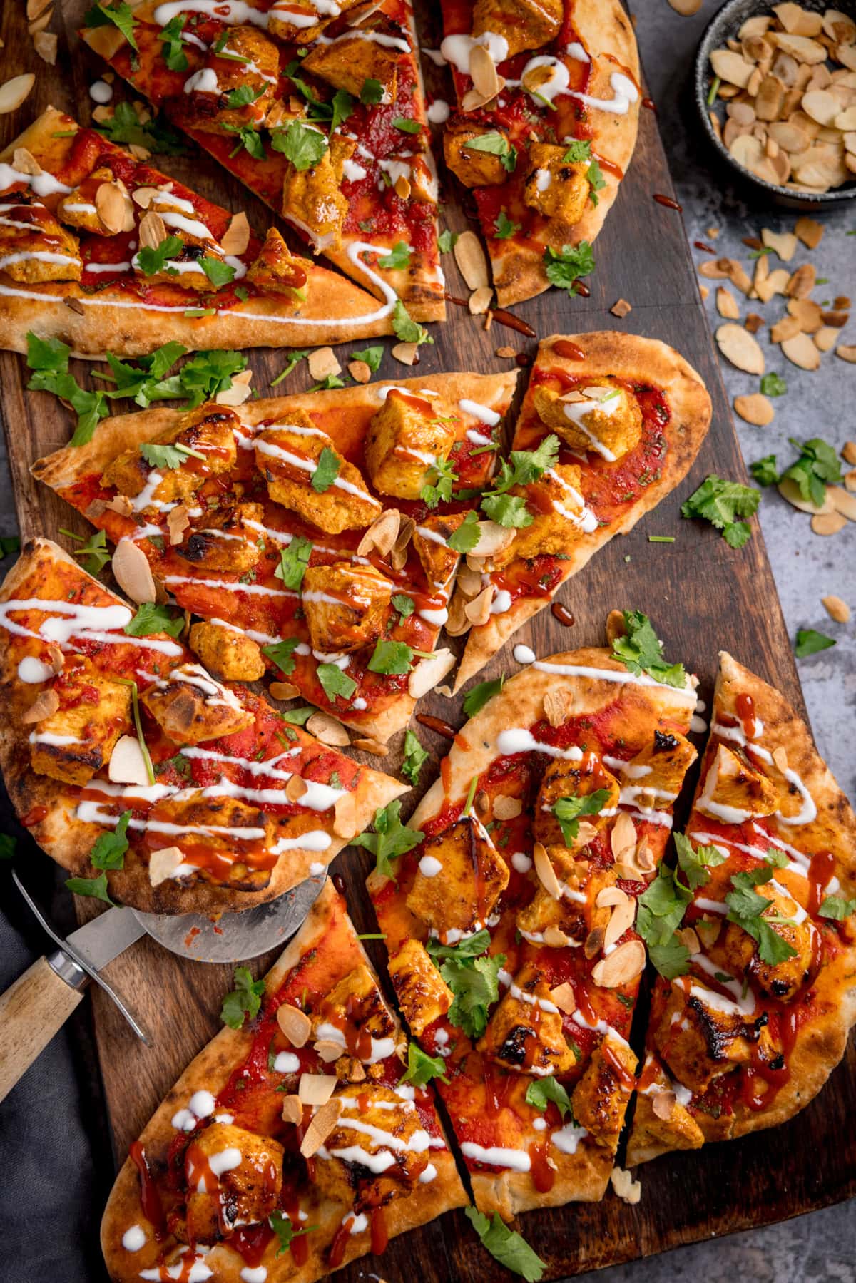 Sliced chicken tikka naan pizza topped with coriander, flaked almonds, and drizzled with sauces on a wooden board. A bowl of flaked almonds sits nearby.