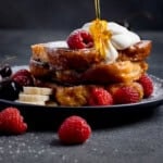 A stack of brioche French toast topped with whipped cream, raspberries, banana slices, and cherries, with syrup being poured over the top.