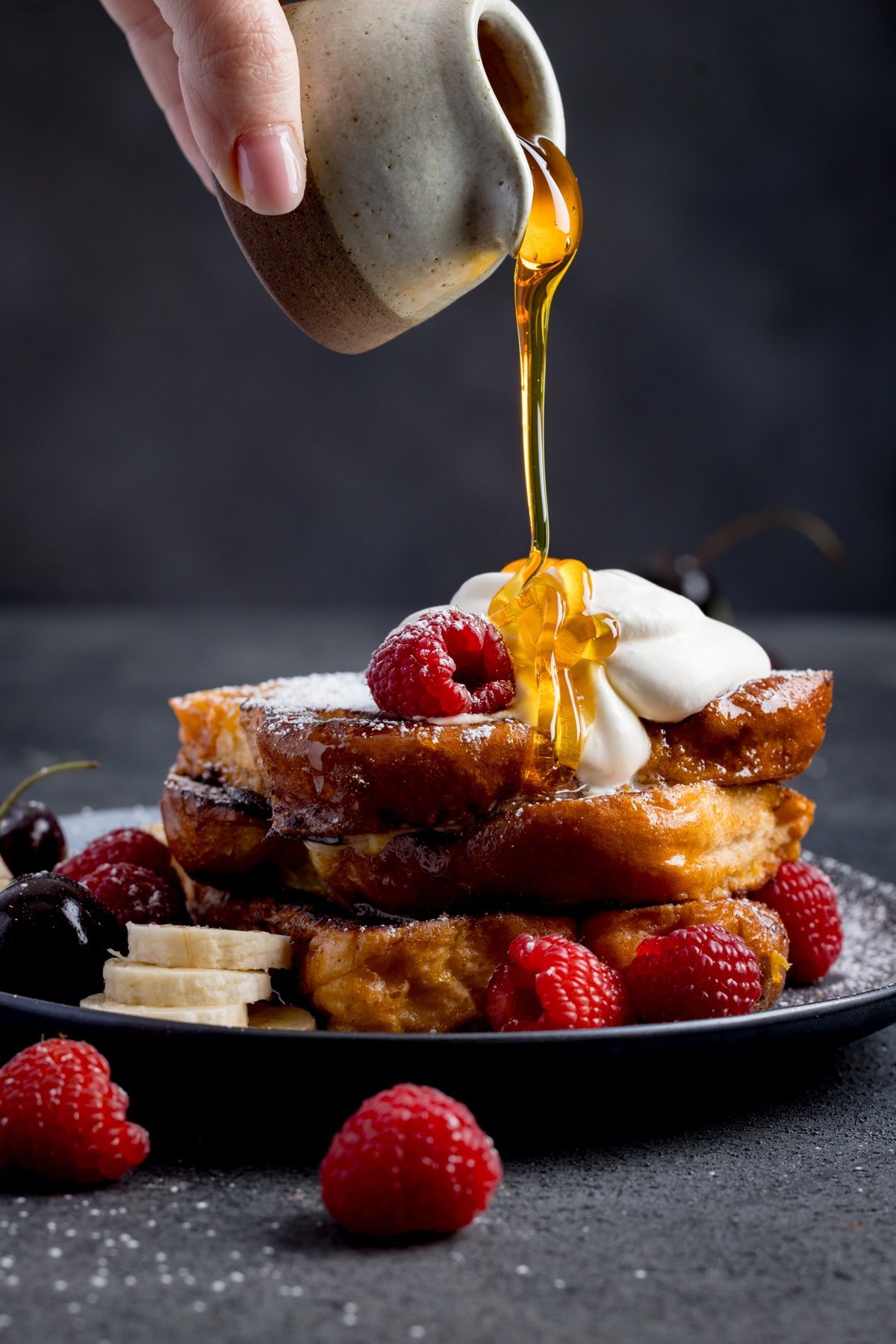 A hand pours syrup onto a stack of caramelised French toast topped with whipped cream, raspberries, cherries, and banana slices on a black plate.