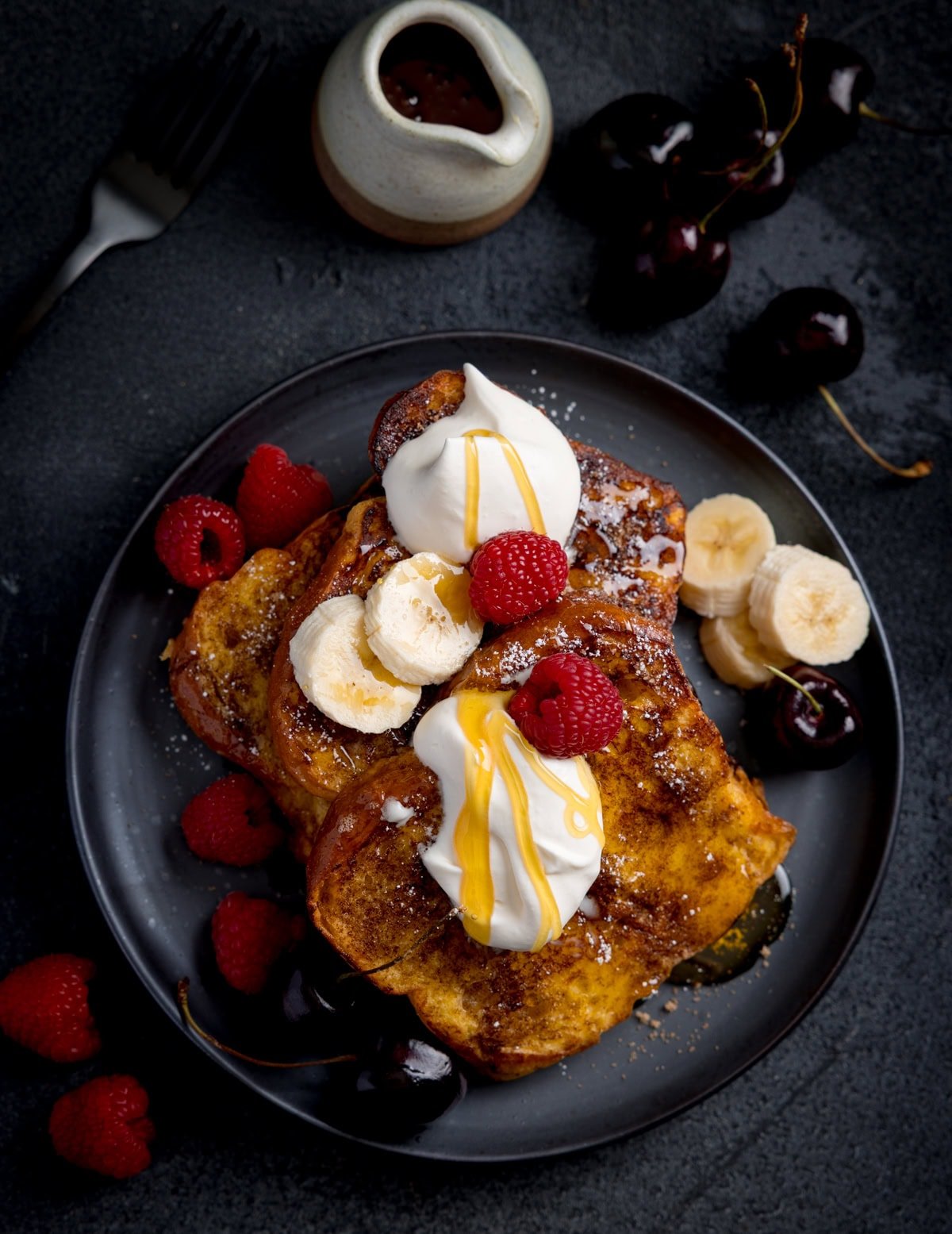 A plate of brioche French toast topped with whipped cream, banana slices, raspberries, cherries, and syrup, with extra fruit and a small jug of syrup nearby.