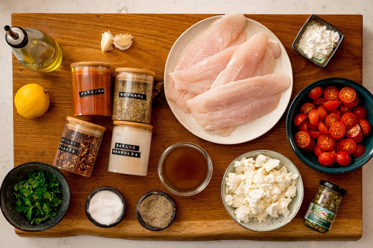 A wooden board displays ingredients for Baked Fish with Lemon and Feta—raw fish fillets, cherry tomatoes, feta cheese, parsley, seasonings, olive oil, lemon, garlic, and spices.
