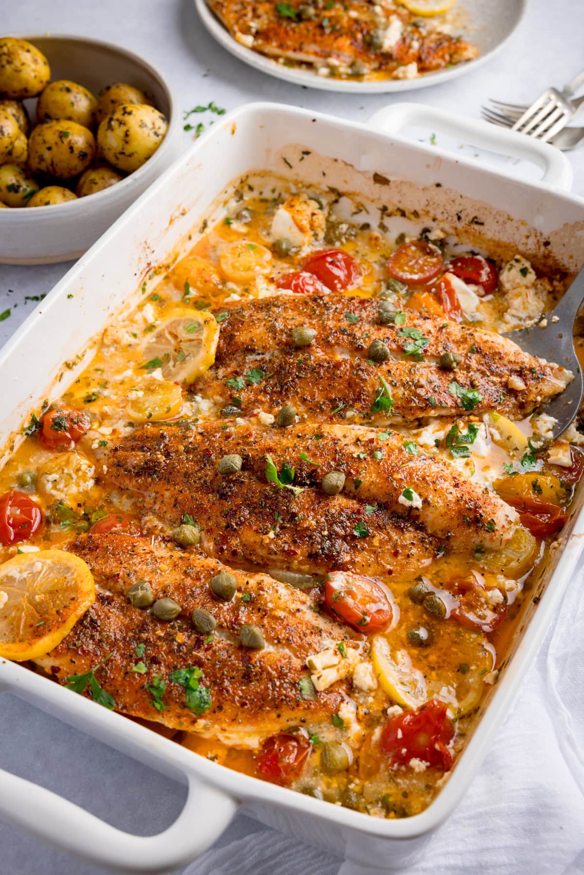 Seasoned fish fillets baked with cherry tomatoes, lemon slices, capers, and herbs in a white oven dish. A bowl of potatoes and a plate sit in the background.