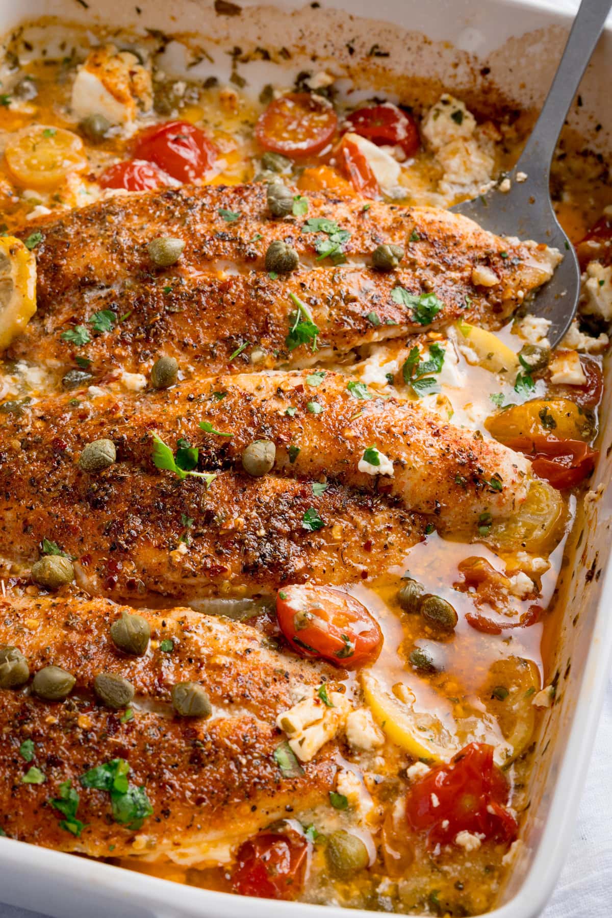 Baked Fish with Lemon and Feta fillets topped with cherry tomatoes, capers, herbs, and sauce in a white baking dish. A serving spoon is nestled in the dish.