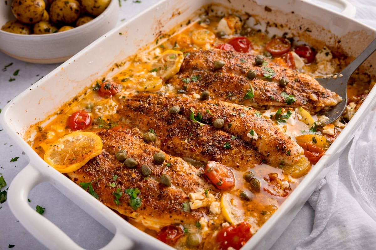 Baked fish fillets in a creamy sauce with capers, lemon slices, cherry tomatoes, and herbs, served in a white casserole dish. A bowl of potatoes can be seen nearby.