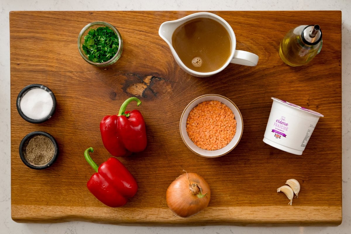 A wooden board displays ingredients for a red lentil pasta sauce: red peppers, an onion, garlic cloves, a bowl of red lentils, creme fraiche, stock, chopped herbs, olive oil, salt and pepper.