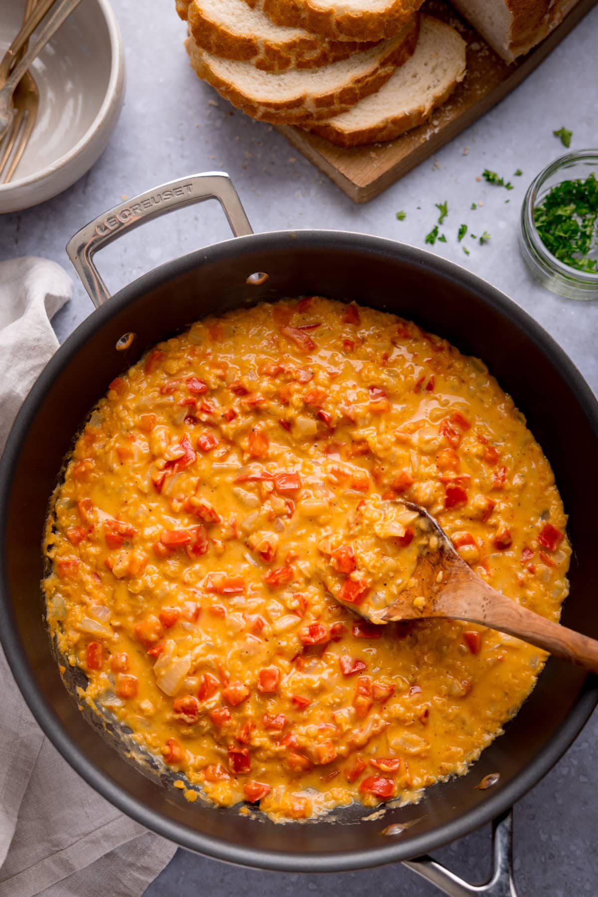 A frying pan filled with a creamy, lentil and red pepper sauce, with a wooden spoon inside. Sliced bread, herbs, and bowls are visible in the background.