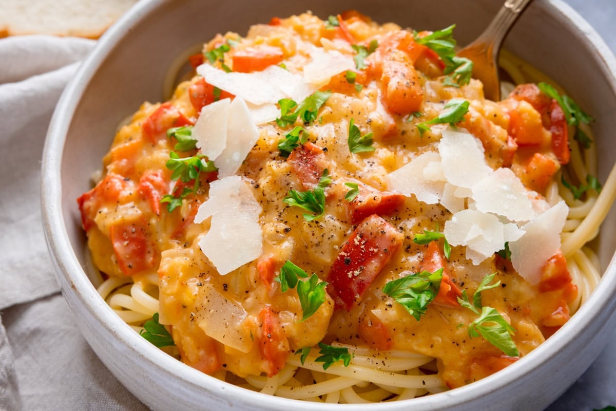 A white bowl of pasta topped with a thick red bell pepper & lentil sauce topped with shaved parmesan