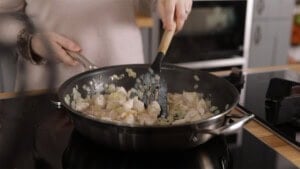 A person stirs diced chicken and chopped onions in a frying pan on a hob.