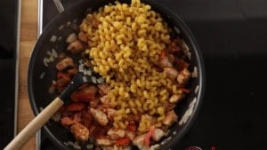 A frying pan on a hob contains chopped onions, diced chicken, red pepper, and uncooked pasta with a spoon resting inside.