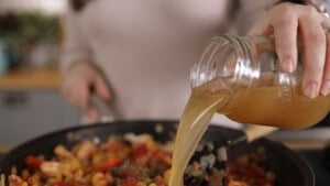 A person pours stock from a glass jar into a pan filled with pasta, chicken and vegetables.