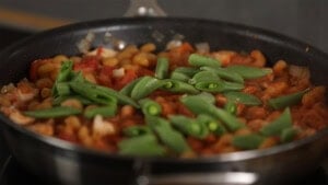 A close-up of a frying pan with pasta in tomato sauce, chopped onions, and fresh green sugarsnap peas on top, cooking on a hob.