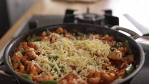 A frying pan on a hob contains pasta with tomato sauce, pieces of chicken, green vegetables, and a topping of grated cheese.