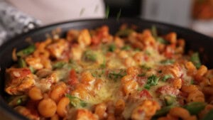 A close-up of a frying pan containing pasta with tomato sauce, melted cheese, chicken pieces, green vegetables, and fresh herbs sprinkled on top.