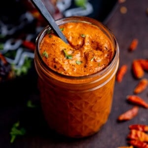 A glass jar filled with thick orange Peri Peri Sauce, garnished with herbs and black pepper, with a spoon inside. Dried red chillies are scattered on the table nearby.