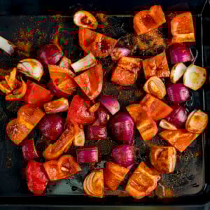 Chopped red peppers and red onions coated with spices on a dark baking tray.