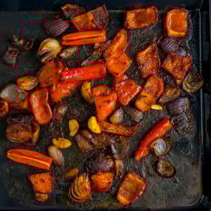 Roasted vegetables, including seasoned red peppers and red onion on a dark baking tray.