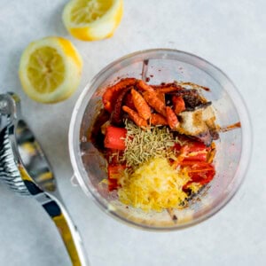 Overhead view of a blender jug on a light surface filled with spices, dried herbs, chillies, and lemon zest for Peri Peri Sauce. Cut lemons and a metal citrus squeezer is next to the the blender jug.