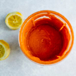 A top-down view of a glass filled with a dark liquid, next to two sliced lemons and a hint of Peri Peri Sauce on a light-coloured surface.