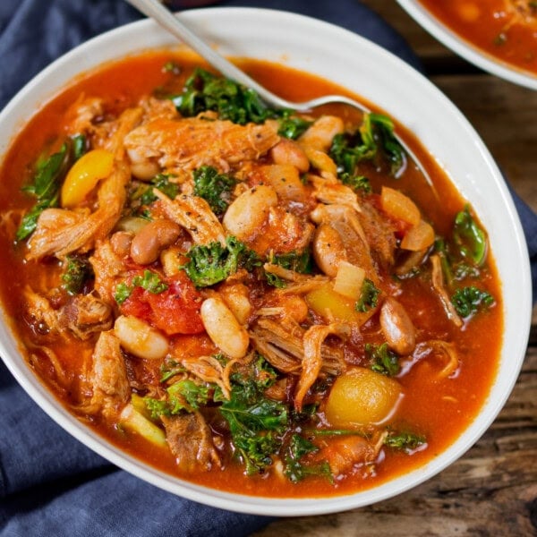 A bowl of pulled pork and bean soup with tender shredded meat, beans, tomatoes, leafy greens, and vegetables in a rich red broth, with a spoon inside the bowl.