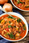A white bowl of hearty pulled pork and bean soup with kale and vegetables, served in a rich tomato-based stew, with a spoon and onions in the background.