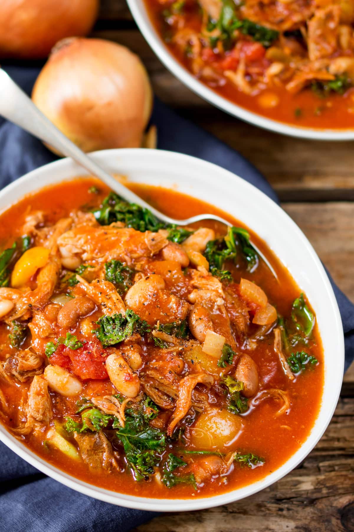 A white bowl of hearty pulled pork and bean soup with kale and vegetables, served in a rich tomato-based stew, with a spoon and onions in the background.