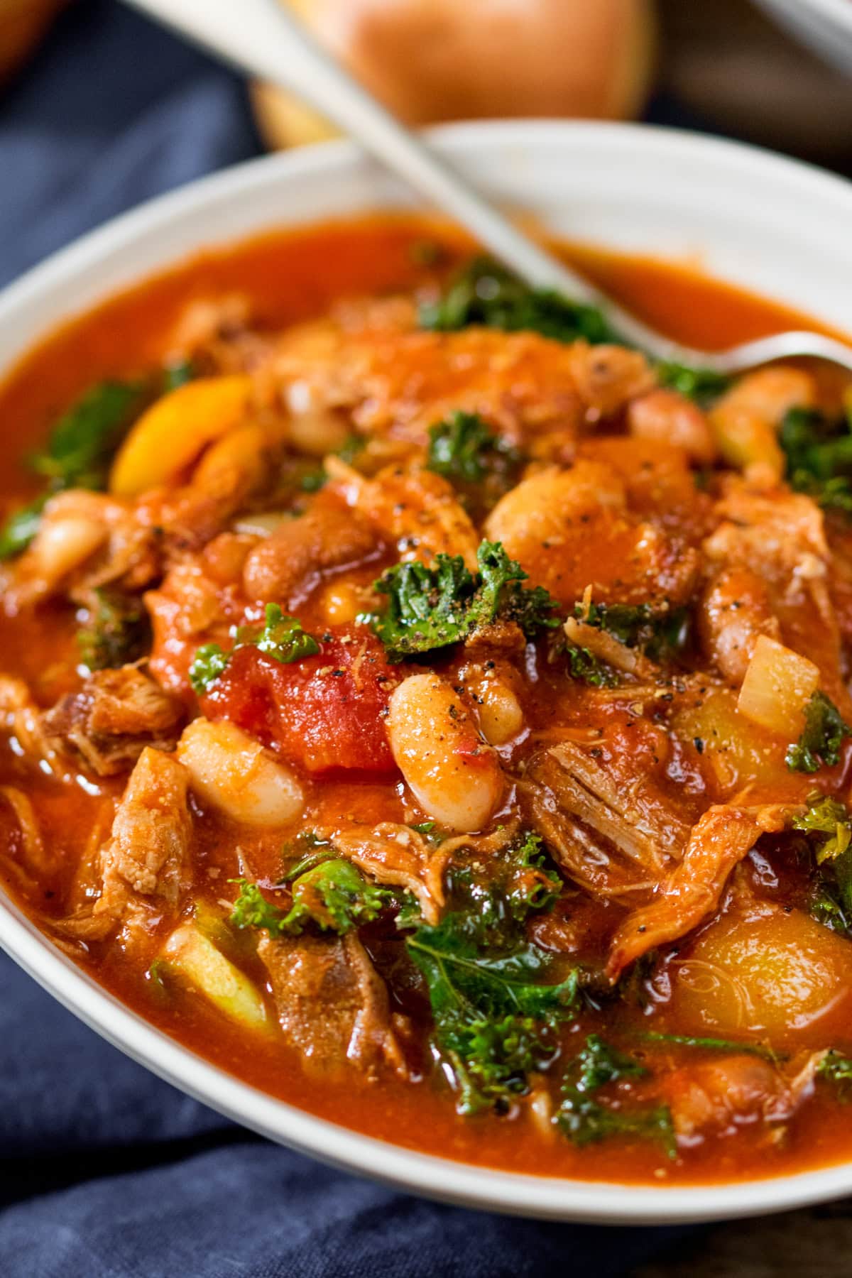 A bowl of hearty pulled pork and bean soup with white beans, tomato-based broth, kale, and vegetables, with a spoon resting inside.