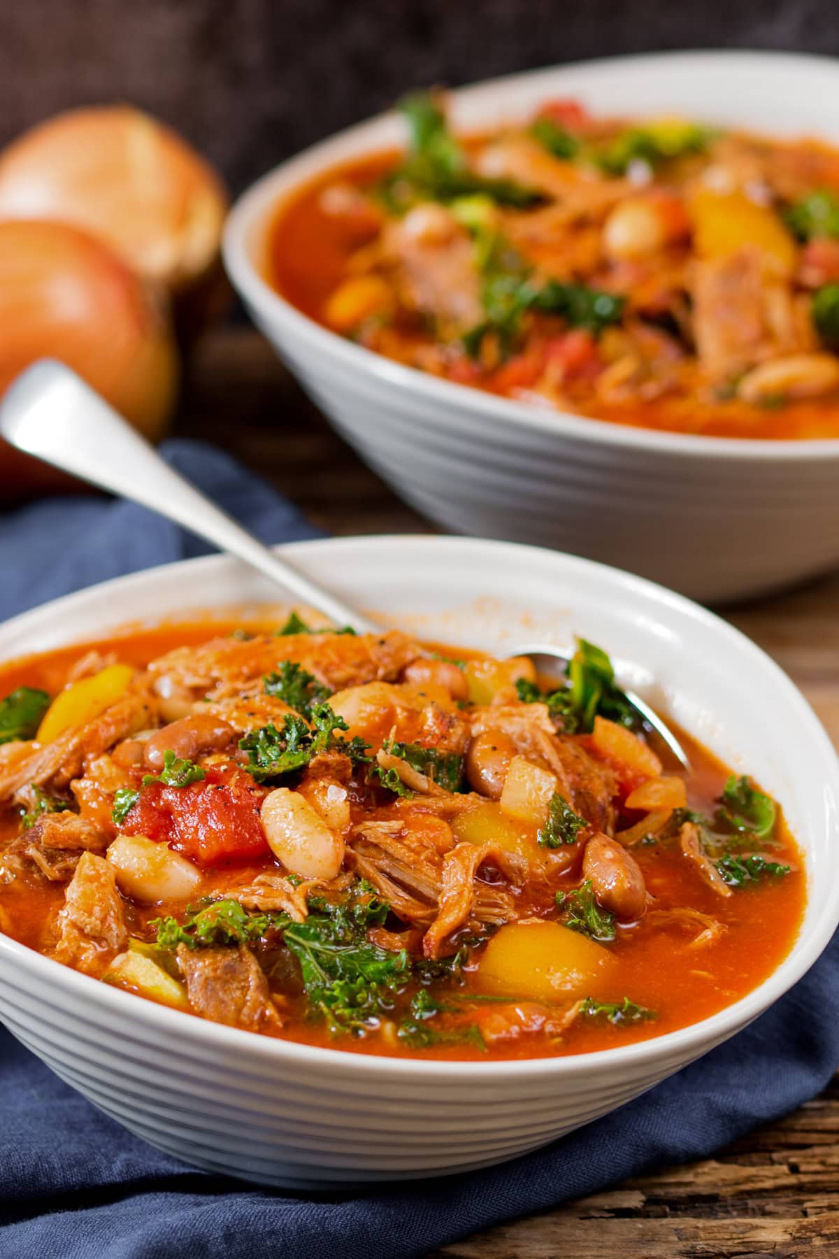Two white bowls filled with hearty pulled pork and bean soup, featuring shredded meat, kale, tomatoes, and stock, with a spoon resting in the front bowl and 2 onions in the background slightly out of focus.