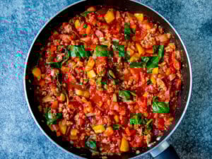 A frying pan filled with a cooked mixture of sausage meat, tomatoes, spinach, onions, and peppers on a blue textured surface.