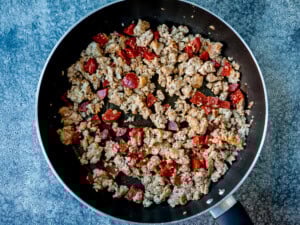 Ground sausage and diced chorizo cooking in a black frying pan on a blue textured surface.