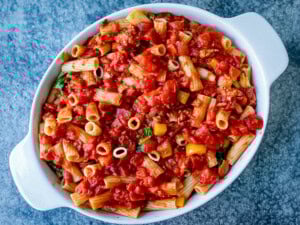 A white oval dish filled with cooked rigatoni pasta mixed with tomato sauce, chopped vegetables, and herbs on a blue textured surface.