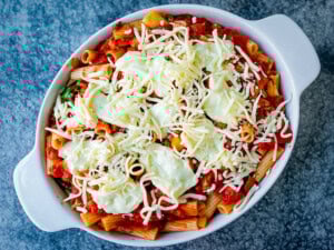 A white oven dish filled with rigatoni pasta, tomato sauce, dollops of brie cheese, and grated mozzarella on top, ready to be baked.