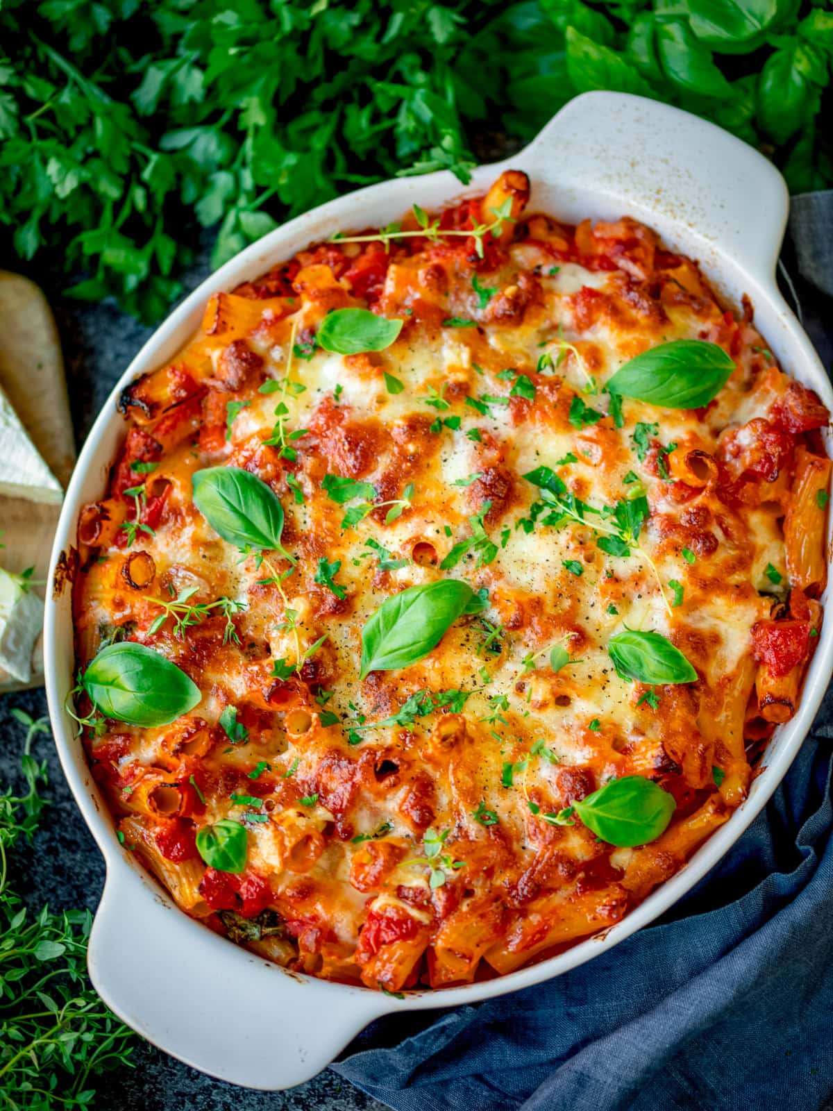 Sausage pasta bake topped with melted cheese, fresh basil, and herbs in a white oval casserole dish on a dark surface.