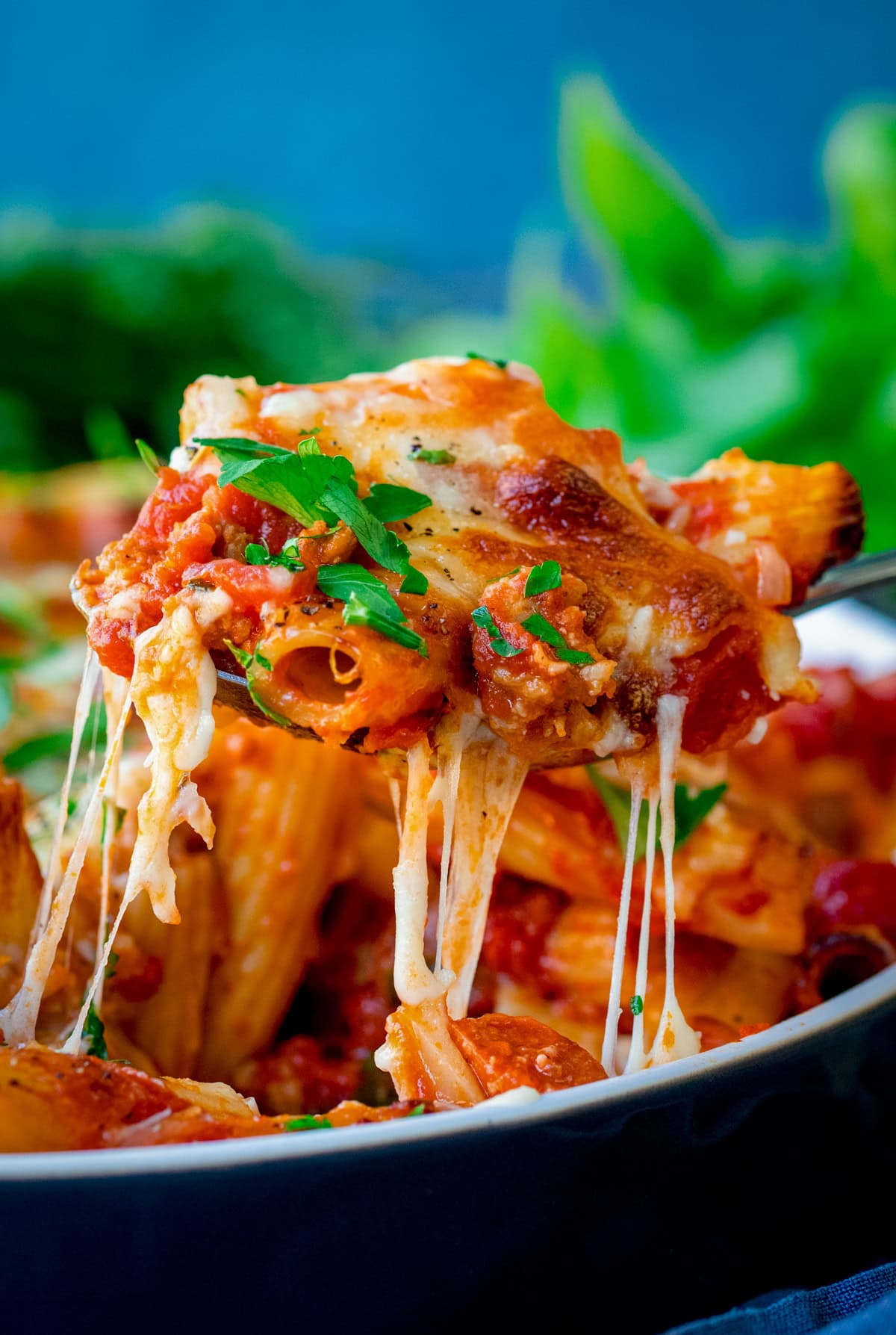 A serving spoon of Sausage Pasta Bake with tomato sauce, melted cheese, and herbs is being lifted from a dish, with gooey cheese stretching from the dish.
