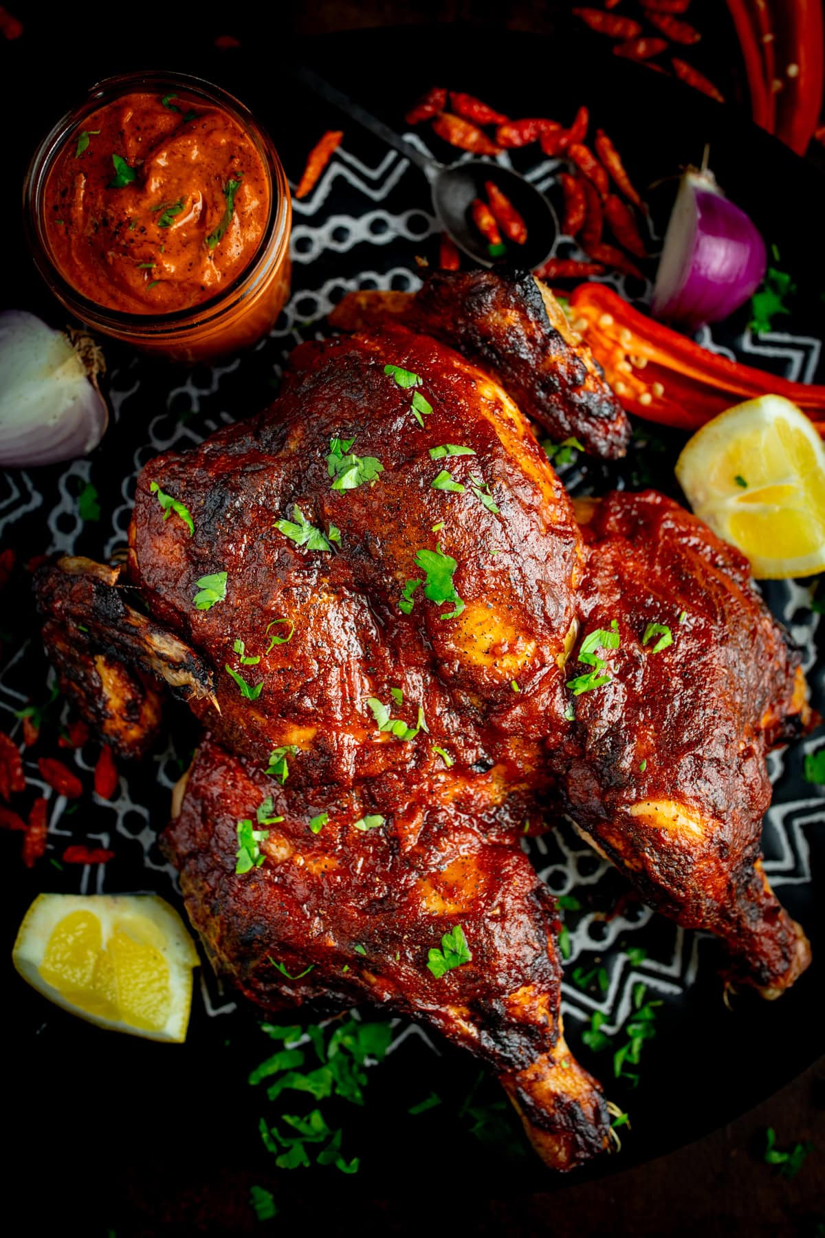 Peri Peri butterflied chicken garnished with chopped herbs, served with lemon wedges, red onion, chilli peppers, and a jar of peri peri sauce on dark serving plate