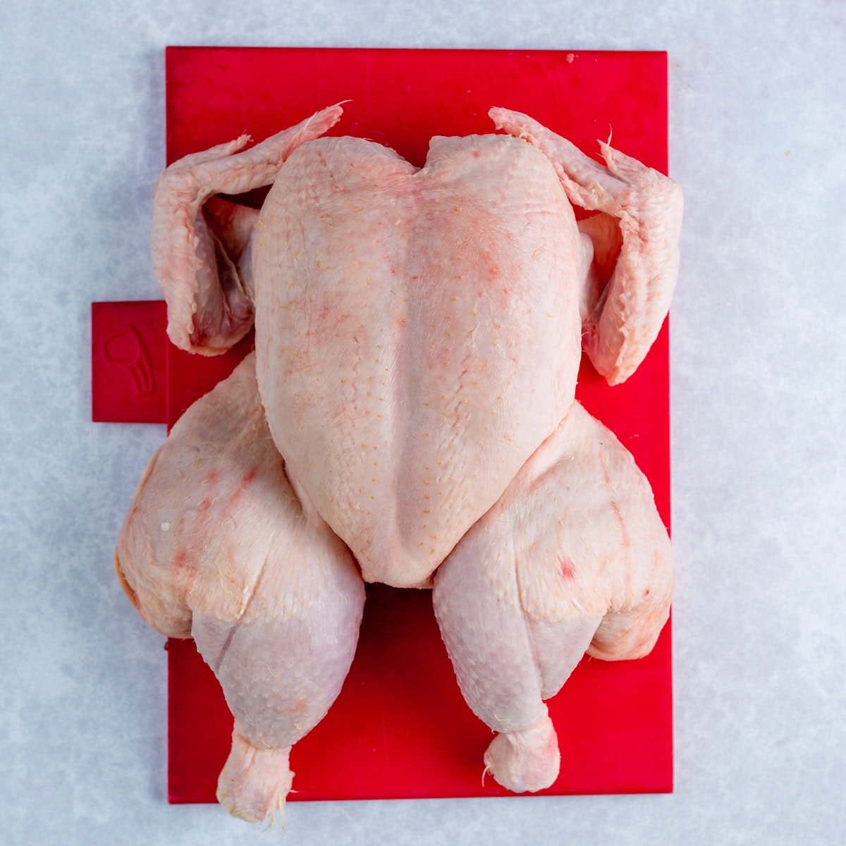 A raw whole spatchcocked chicken lying breast-side-up on a red chopping board against a light grey background.