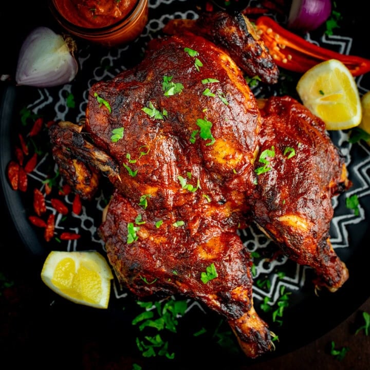 Square image of Peri Peri butterflied chicken garnished with chopped herbs, served with lemon wedges, red onion, chilli peppers, and a jar of peri peri sauce on dark serving plate