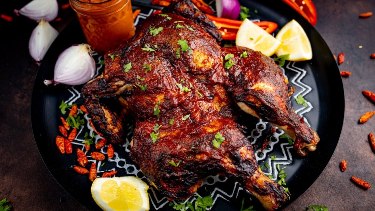 A butterflied chicken, roasted whole and garnished with herbs, lemon wedges, and red chilli peppers, is served with a jar of sauce on a black plate.