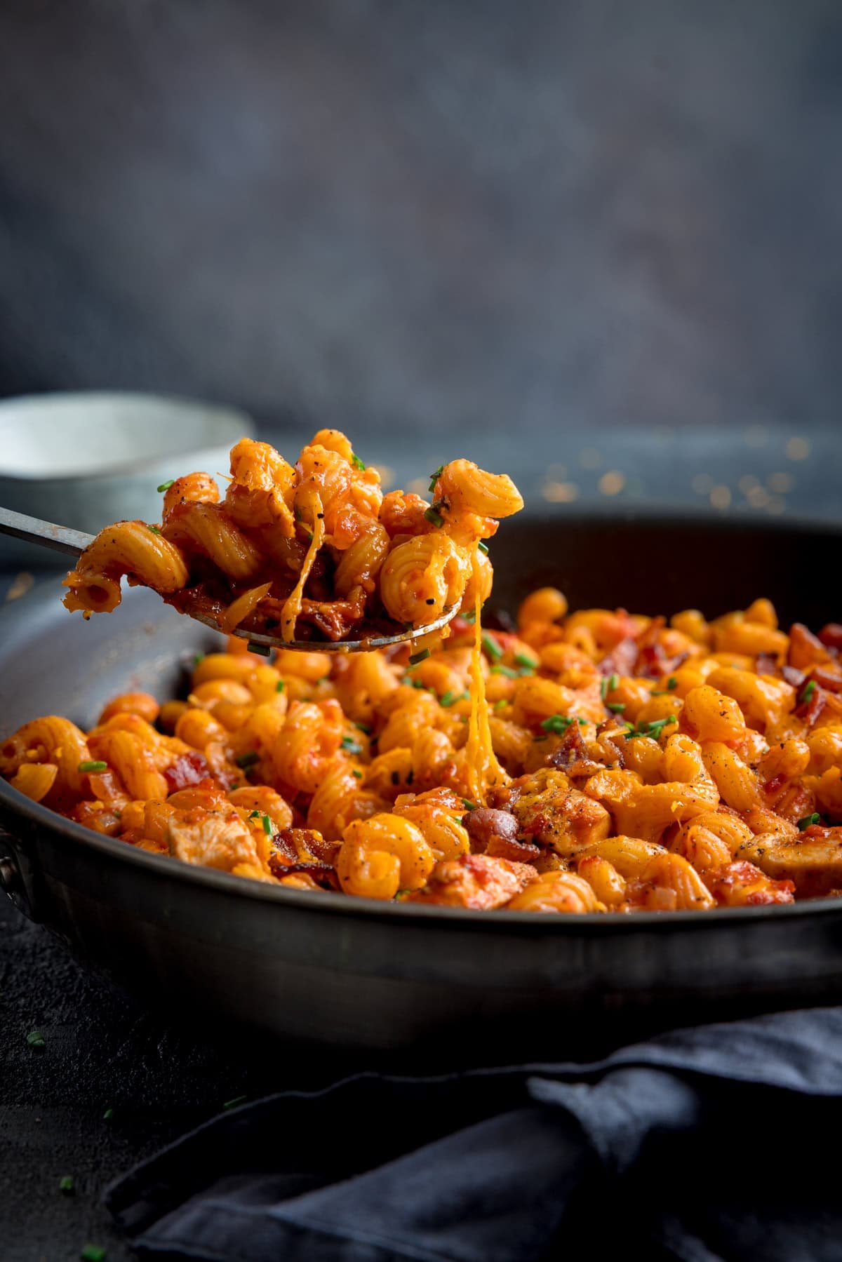 A frying pan filled with cheesy BBQ Chicken Pasta—corkscrew pasta in tangy BBQ tomato sauce, topped with melted cheese, herbs and chicken. A spoon is lifting a serving of the pasta.