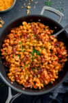 A frying pan filled with cooked pasta, BBQ chicken, bacon, a rich tomato sauce, and chopped chives. A spoon rests in the pan.
