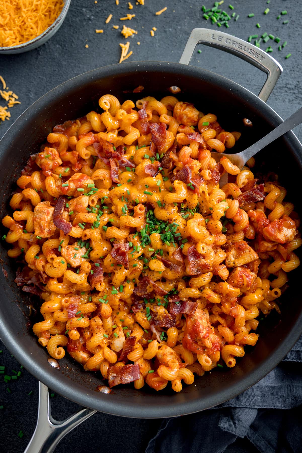A frying pan filled with cooked pasta, BBQ chicken, bacon, a rich tomato sauce, and chopped chives. A spoon rests in the pan.