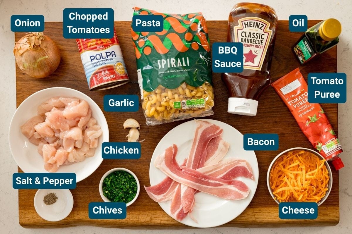 A wooden board displaying labelled ingredients for a cheesy BBQ chicken pasta, including chicken, bacon, pasta, cheese, onion, garlic, chopped tomatoes, BBQ sauce, tomato purée, oil, chives, and salt & pepper.
