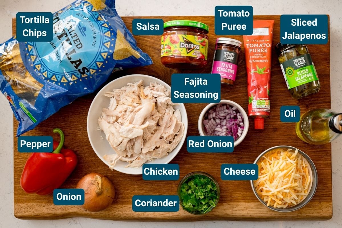 A wooden board holds ingredients for a delicious baked salsa chicken: tortilla chips, salsa, tomato purée, jalapeños, oil, cheese, coriander, shredded chicken, fajita seasoning, red onion, onion and a red pepper.