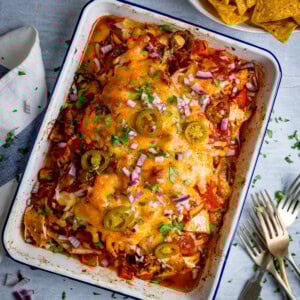 A baked casserole dish filled with tender baked salsa chicken, topped with melted cheese, jalapeños, red onions, and chopped herbs; tortilla crisps sit in a bowl nearby.