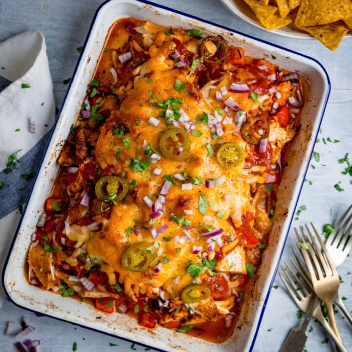 A baked casserole dish filled with tender baked salsa chicken, topped with melted cheese, jalapeños, red onions, and chopped herbs; tortilla crisps sit in a bowl nearby.